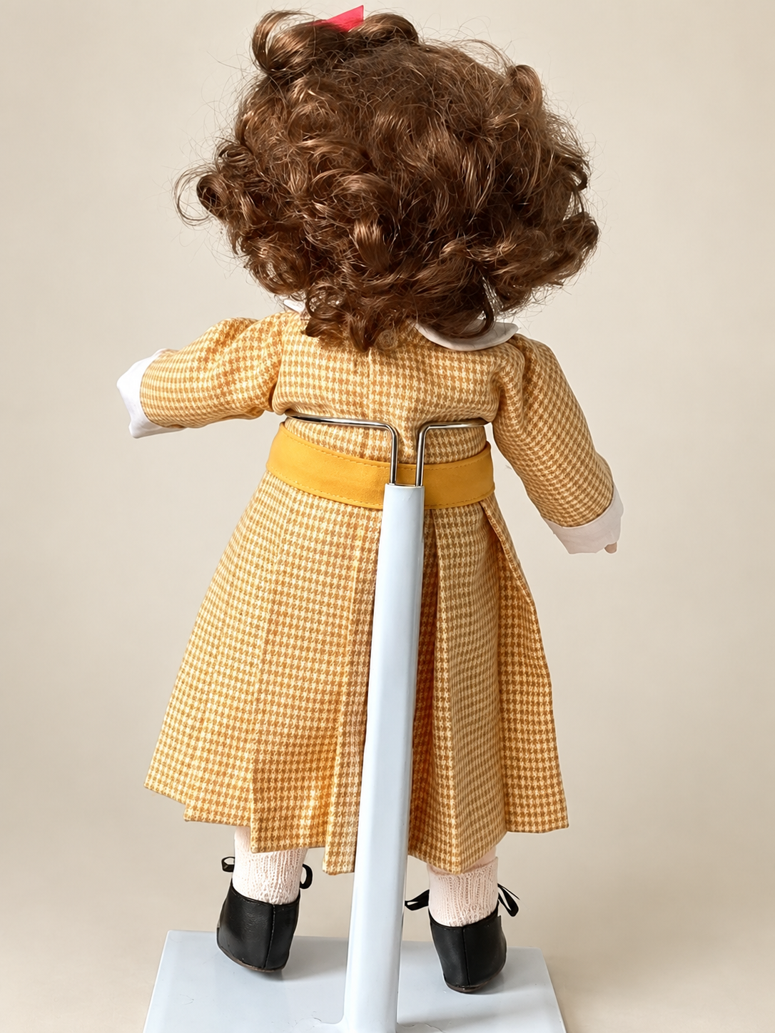 Une tête retrouvée, une poupée recréée — Femmes d’Aujourd’hui, collection de Françoise — Vue de dos de la poupée de Françoise : boucles brunes, robe à carreaux ocre et blancs, ceinture jaune, chaussettes claires et support de présentation — vue 2.